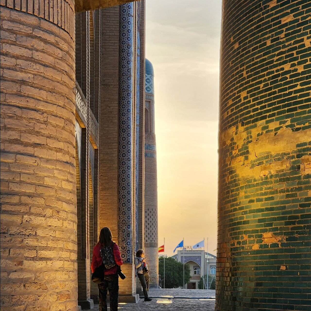 Visitors walk between the tiled Kalta Minor minaret and a madrasa in Khiva at sunset.